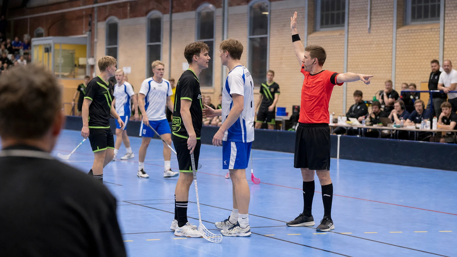Domaren håller upp armen mot utvisningsbåset medan två spelare pratar och lagen samlas bakom dem