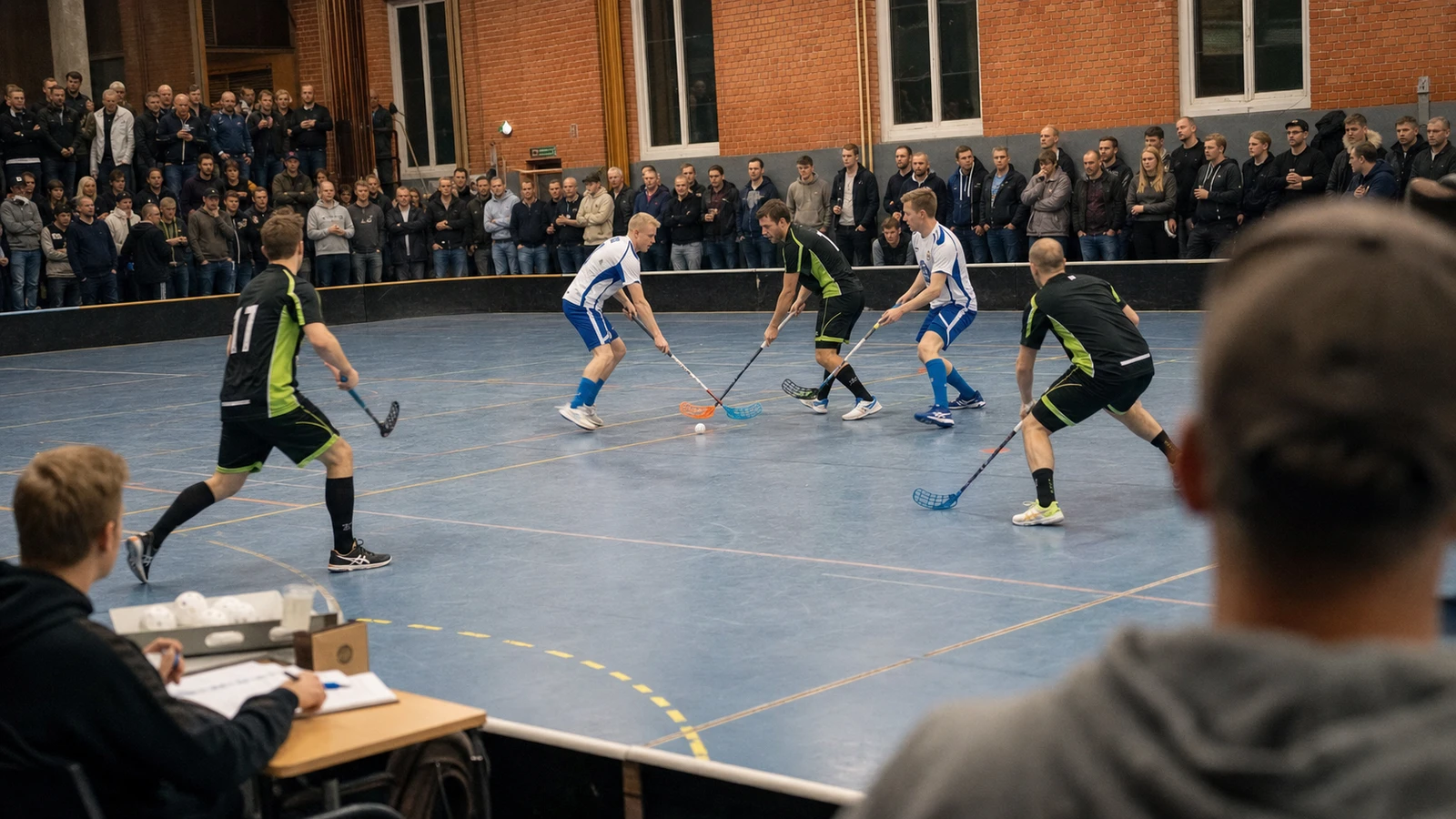 En match i en liten hall där ett fyrtiotal åskådare står tätt längs sargen och en sekretariat sitter vid ett bord